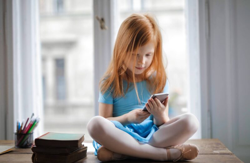 calm small ginger girl sitting on table and using smartphone in light living room